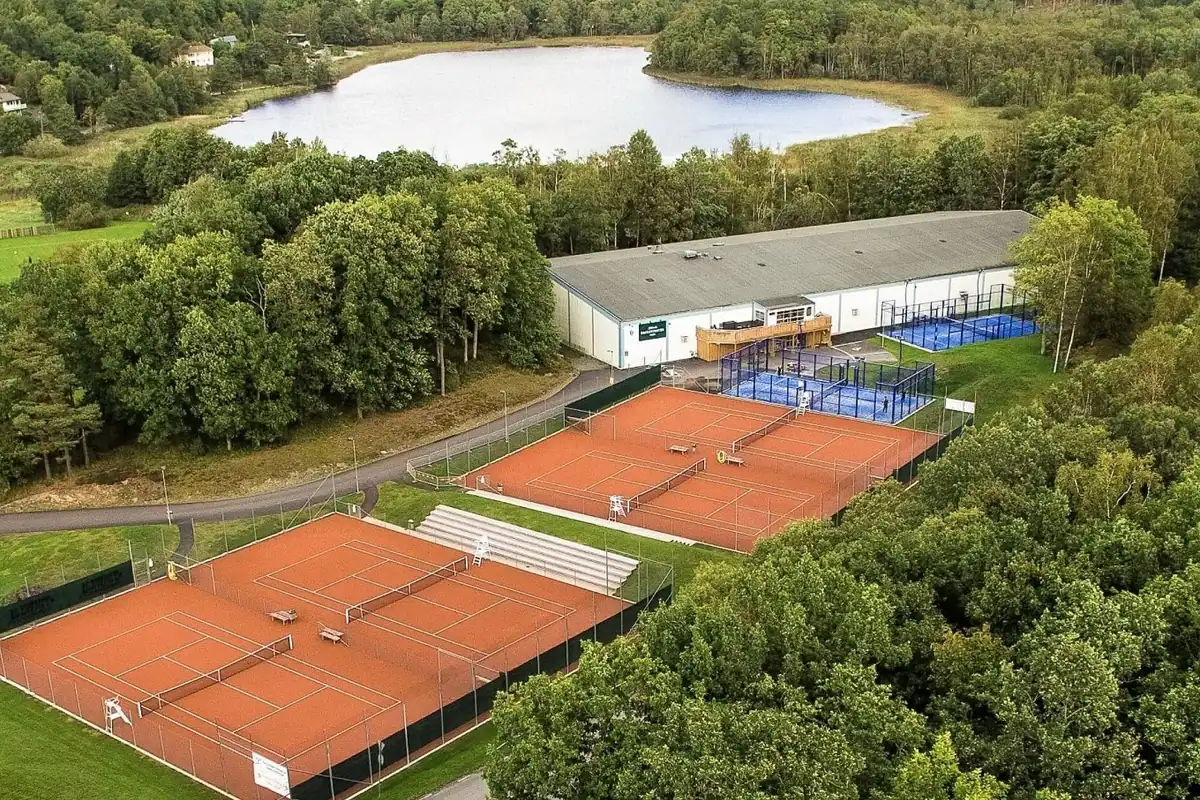 Anläggningen från luften med fyra grusbanor, läktaren, padelbanorna, inomhushallen och sjön bakom