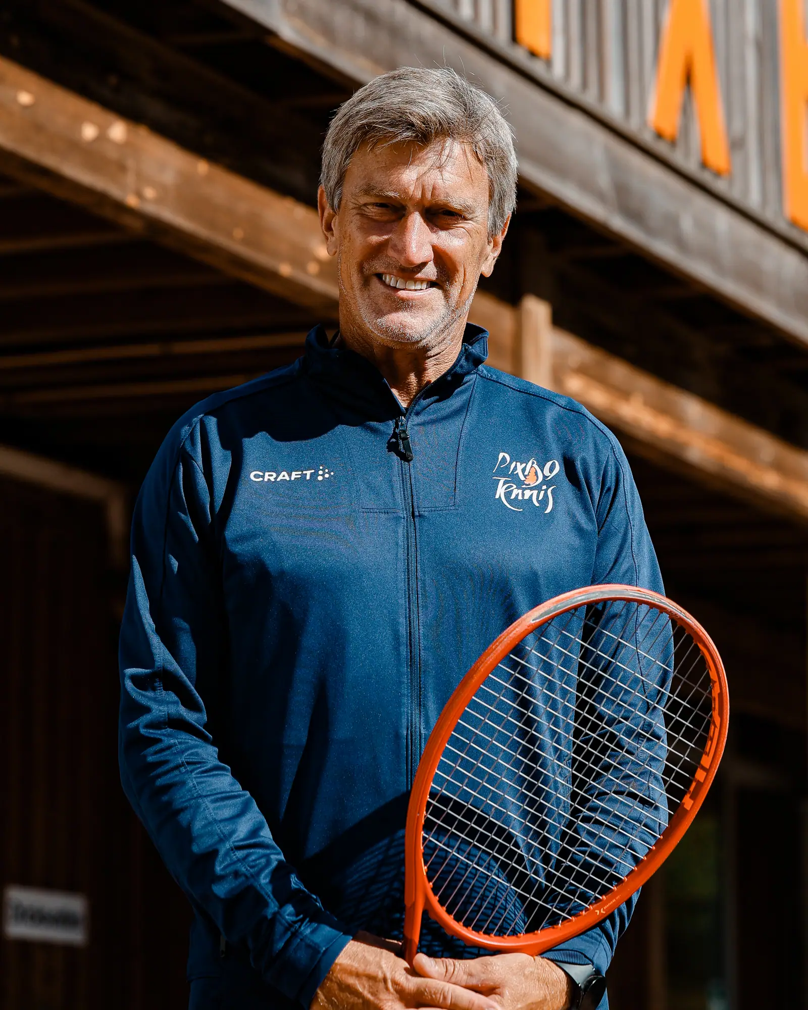 Johan Alvén, tennistränare, med orange racket framför klubbhusets fasad