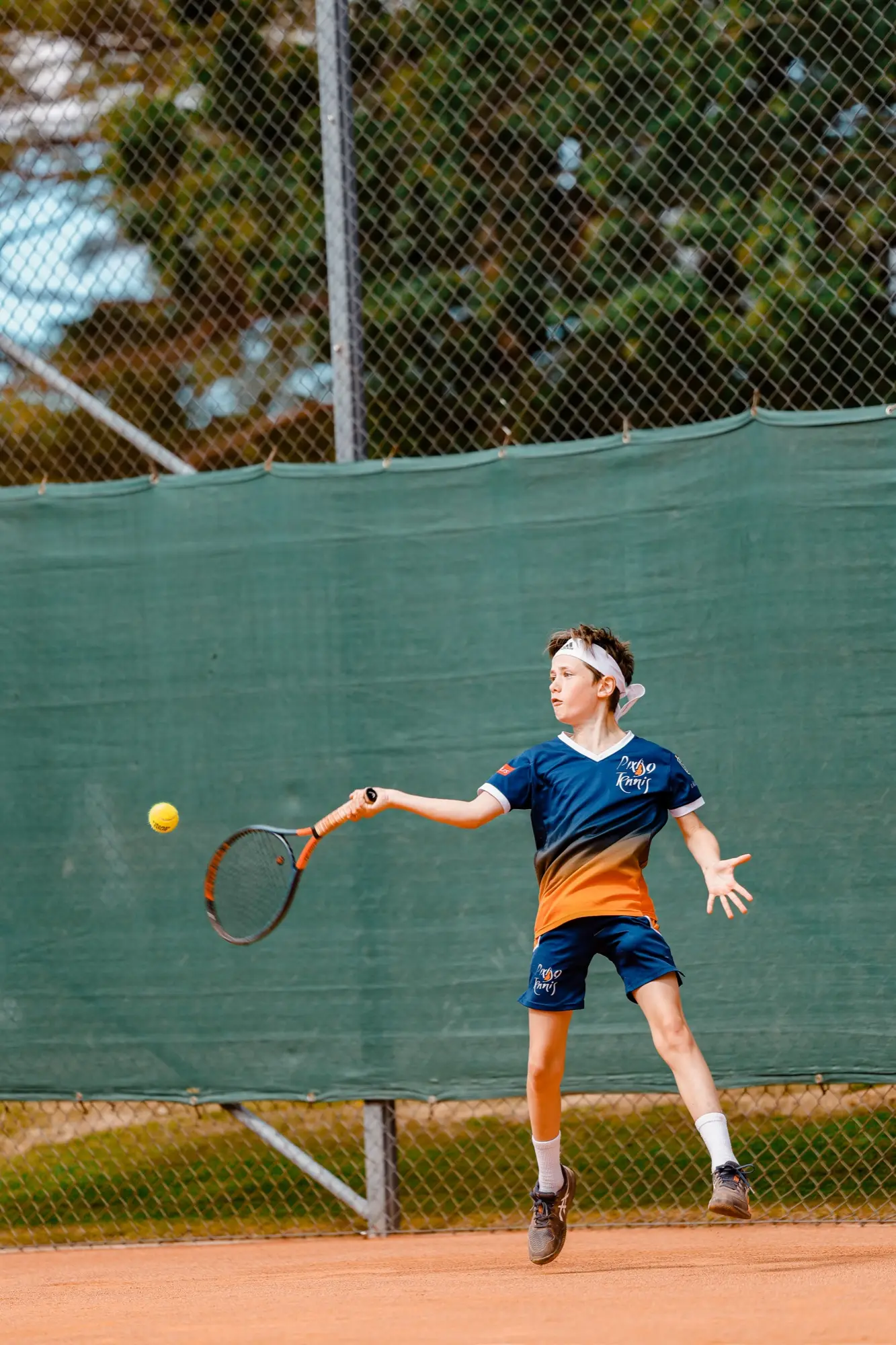 Junior i klubbens tröja slår en forehand med bollen synlig i luften