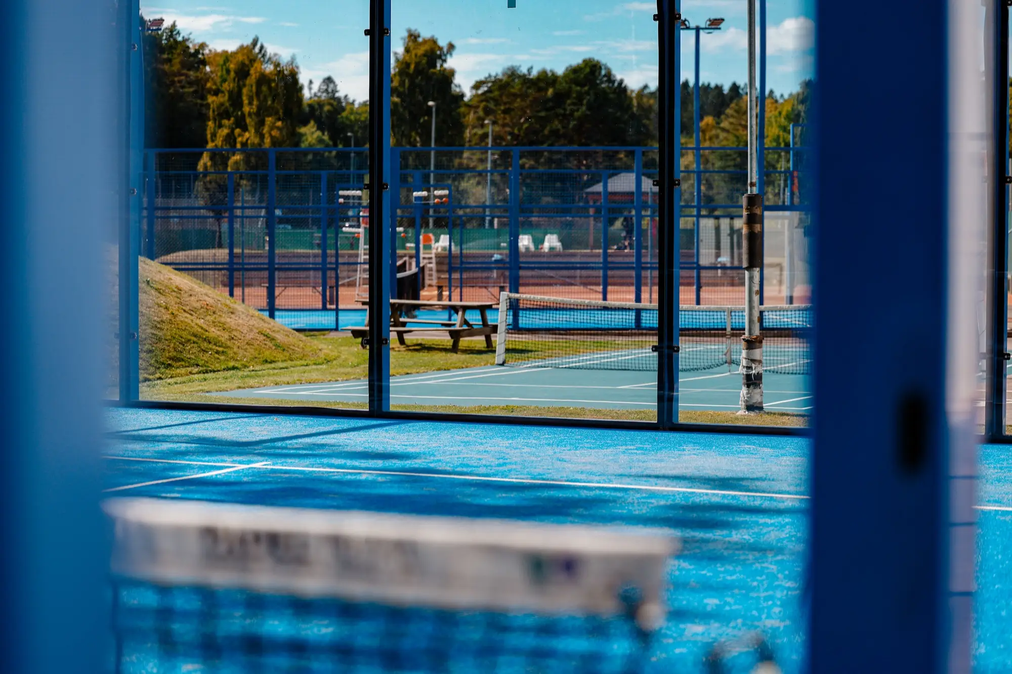 Utsikt från padelbanan genom glasväggen mot den andra banan och grusbanorna