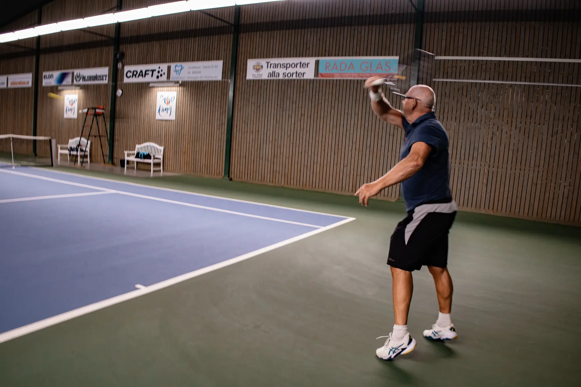 Spelare slår en serve på inomhusbanan under sponsorskyltarna längs väggen