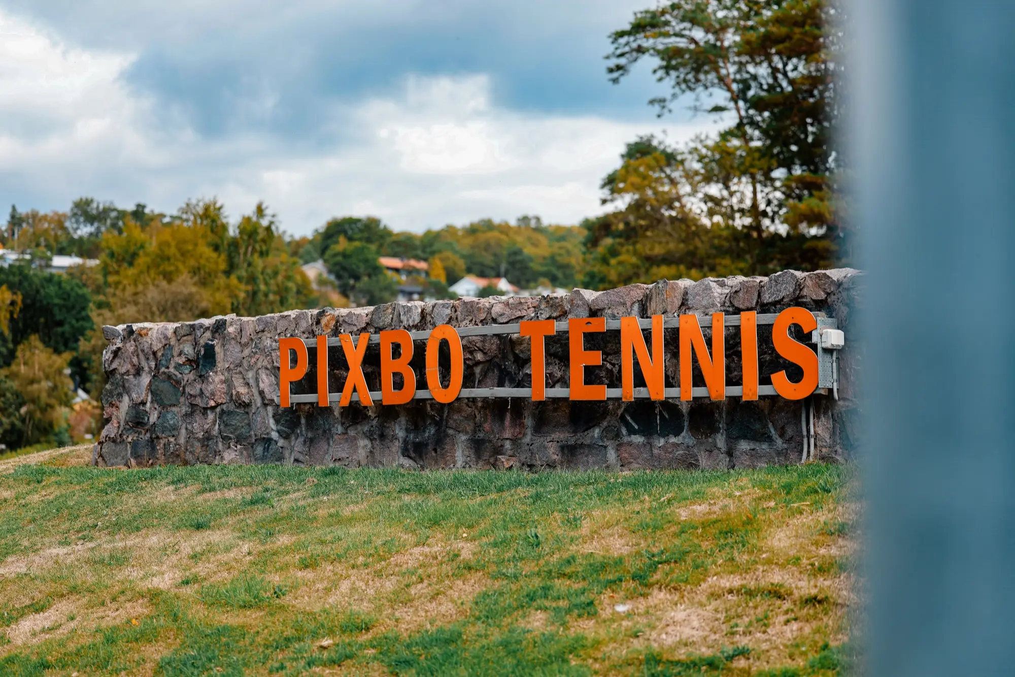 Stenskylten med Pixbo Tennis i orange bokstäver med villabebyggelsen bakom