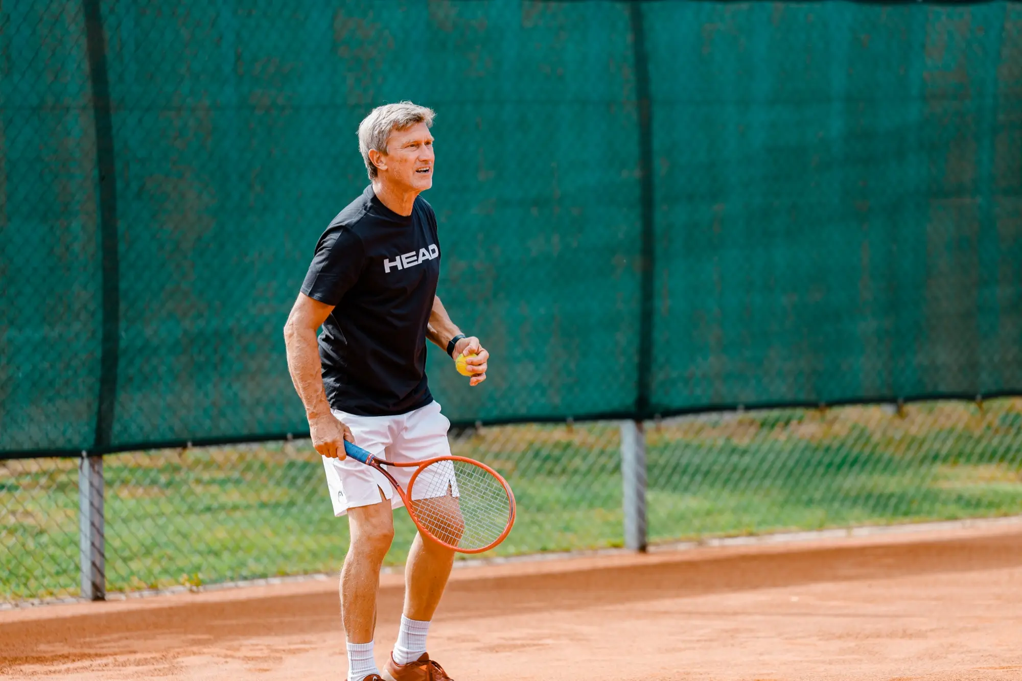 Vuxen spelare med orange racket i slagposition på grusbanan en solig sommardag