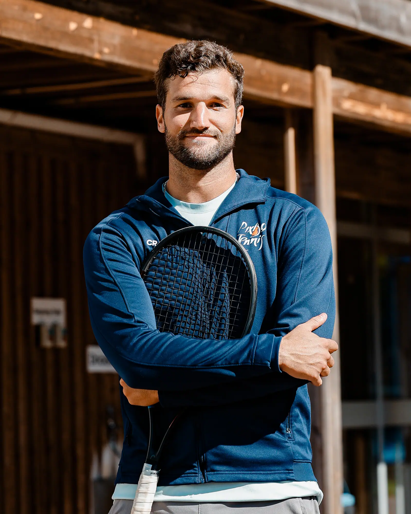 Yannick Trollstad, tennistränare, med racket framför klubbhusets orange fasadtext